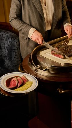 Person slicing rare steak on a carving station with plated steak frites and sauce on a white dish.