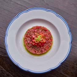 Steak tartare served on a white plate with a blue rim, garnished with finely chopped greens and cubes of vegetables.