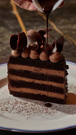 Chocolate layered cake with chocolate mousse and shavings, being drizzled with chocolate sauce on a white plate.