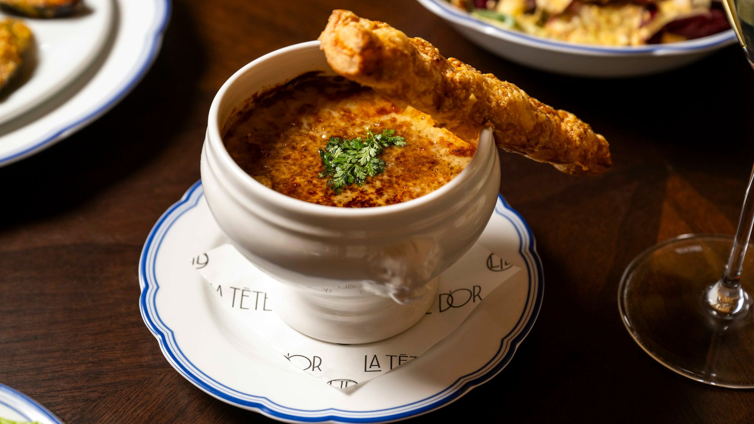 A serving of French onion soup in a white bowl, garnished with herbs and accompanied by a crispy bread stick.