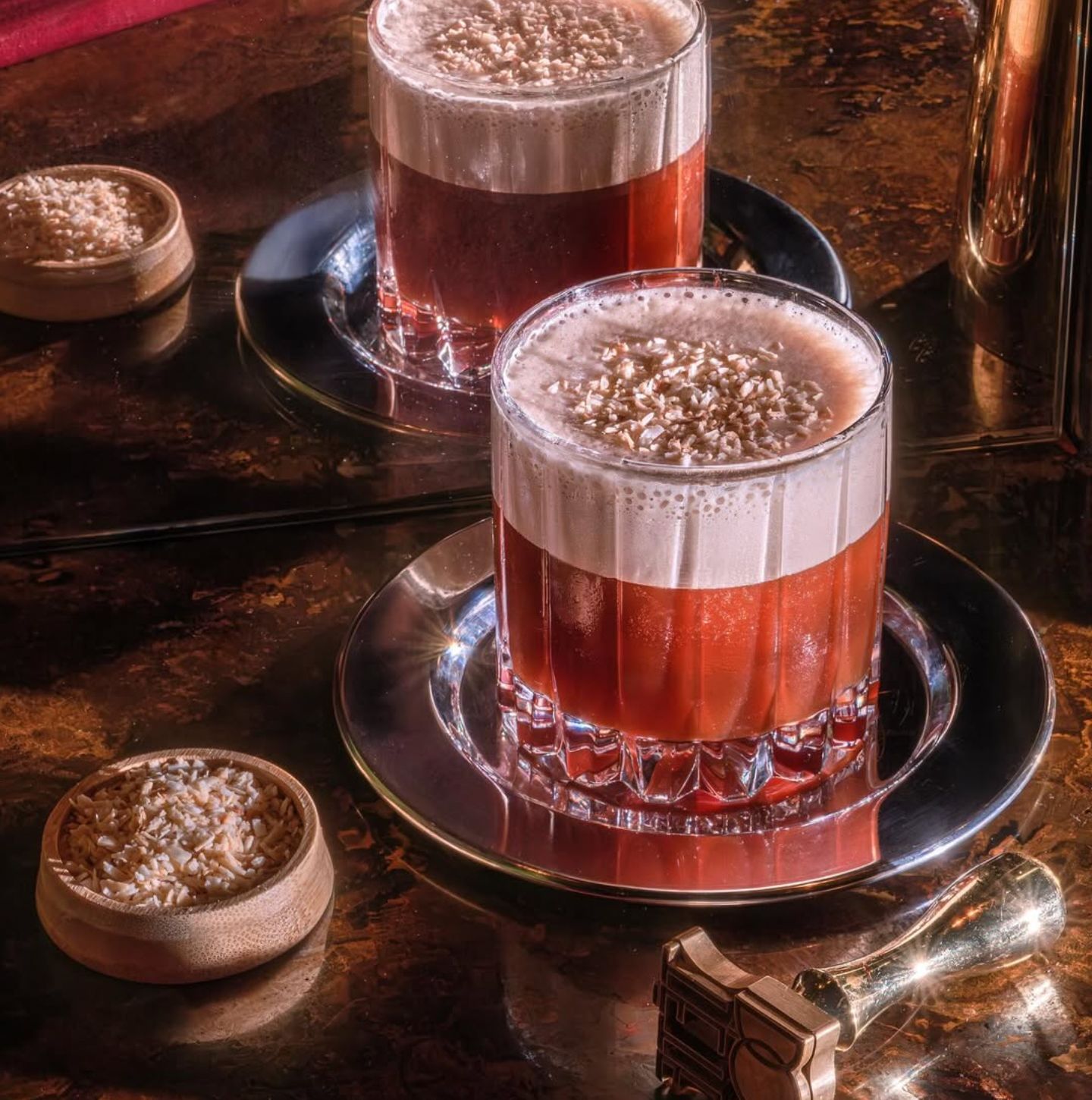 Two cocktails are displayed on a polished surface with garnishes, showcasing a vibrant presentation.